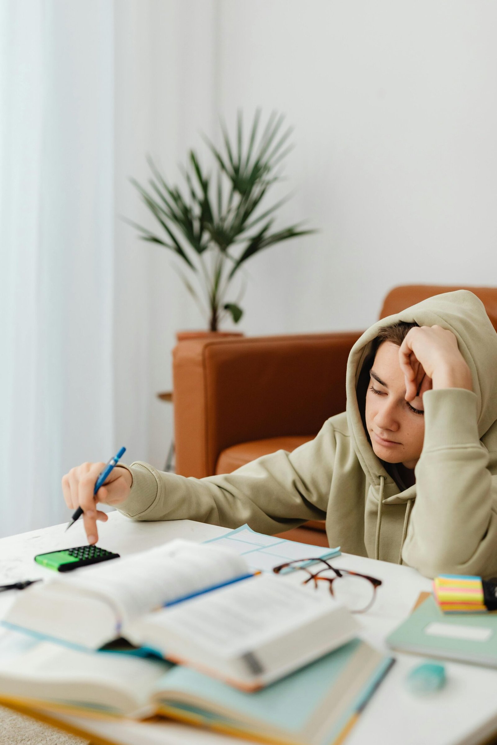 Teenage boy in a hoodie studying at home, looking tired, with books and a calculator.
